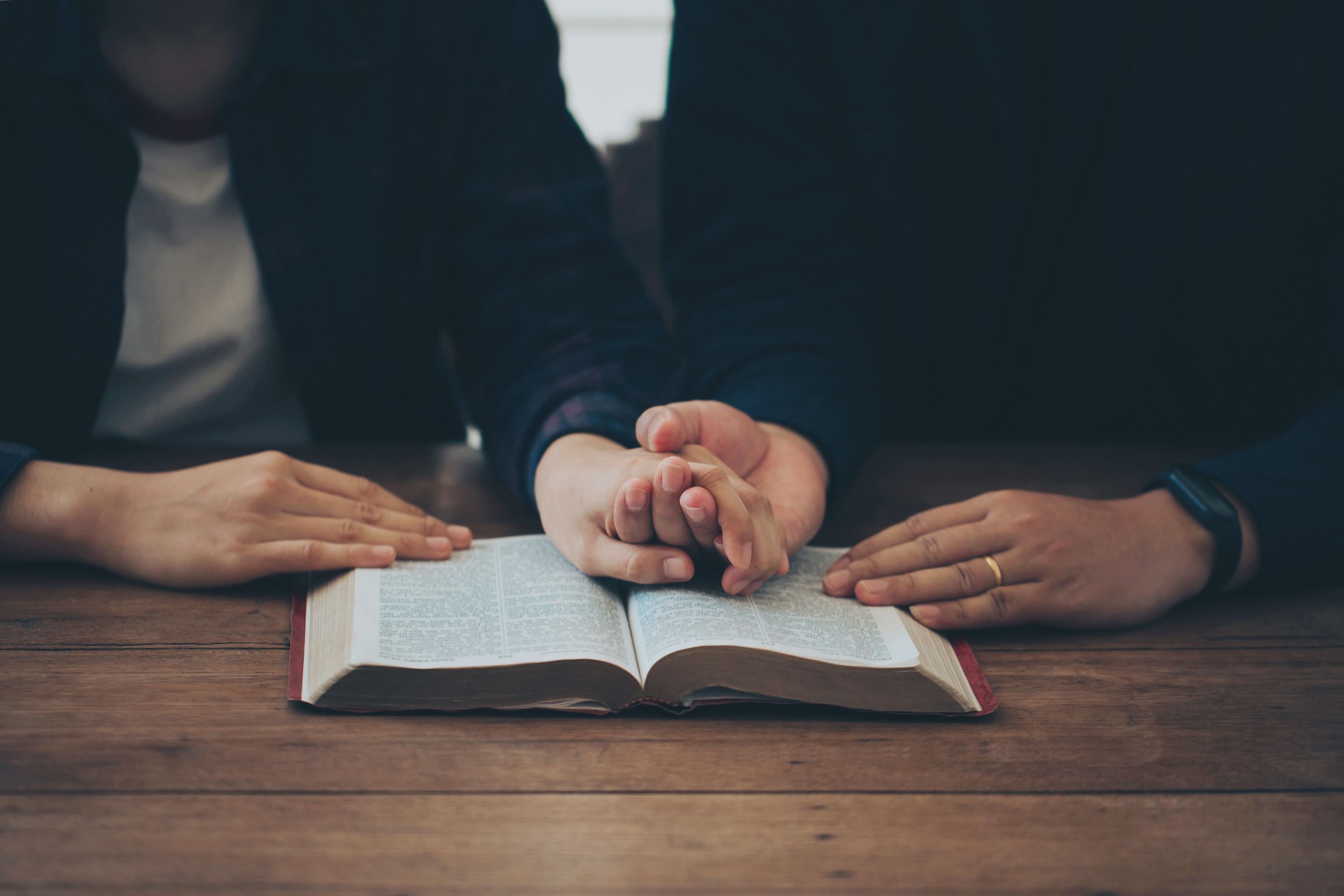 Two Christian couples holding each other's hands praying together over the bible on a wooden table. Begging for forgiveness and believing in goodness. Christian life crisis prayer to god.